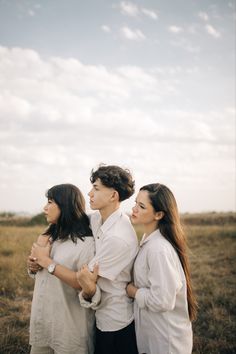 three people standing together in a field with grass and clouds behind them, one woman is hugging the other