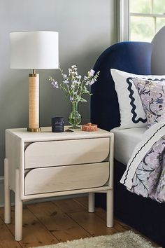 a nightstand with flowers on it next to a blue chair and white bed in a bedroom