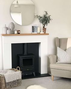 Candles, vases, plants, and books fill the thin wooden mantel in this modern living room. The wooden mantel complements the white fireplace and white background by giving the neutral design a warm accent. A wicker basket filled with blankets adds a rustic charm to the space while providing convenient storage nearby the black wood-burning stove. Add a white and black striped cushion to the taupe armchair for extra comfort...   Image: thebarlowhome_