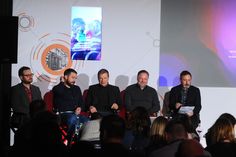 four men are sitting on chairs in front of a crowd and one man is speaking