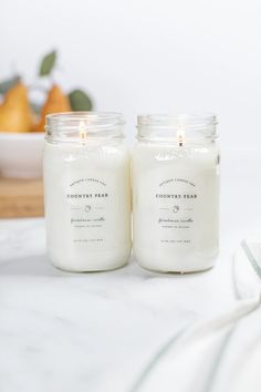 two candles sitting next to each other on top of a white countertop with fruit in the background