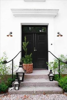 a black door and some plants in front of the steps to a white building with two lights on each side