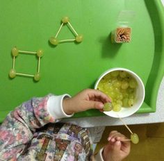 a young child eating grapes from a bowl on a green tray with matchsticks