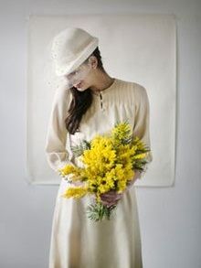 a woman in a white dress and hat holding yellow flowers next to a painting on the wall