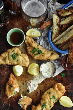 fish and chips on a wooden table next to some beer, sauces and lemon wedges