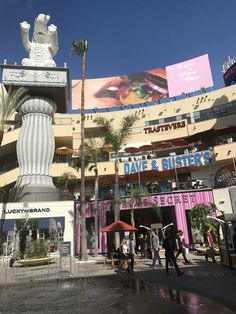 people are walking around in front of a building with palm trees and billboards on it