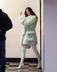 a woman standing in front of a door wearing a green coat and white pants with her hand on her hip