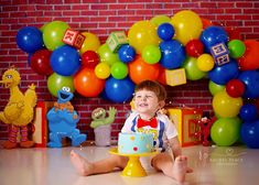 a little boy sitting in front of a cake with balloons on the wall behind him