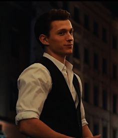 a man wearing a vest and tie standing in front of a tall building at night