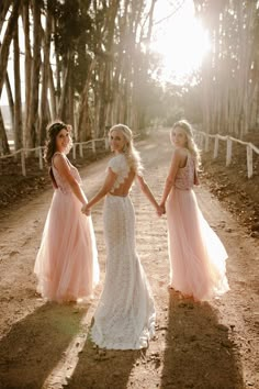 three bridesmaids in pink dresses holding hands on a dirt road with trees behind them