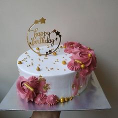 a white and pink birthday cake with gold decorations on it's top is being held by a person