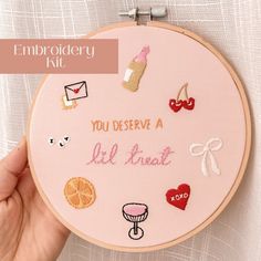 embroidery kit with embroidered words and pictures on the front, in pink hoop held by a person's hand