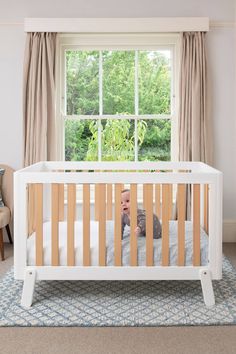 a baby in a white crib looking out the window at trees and bushes outside