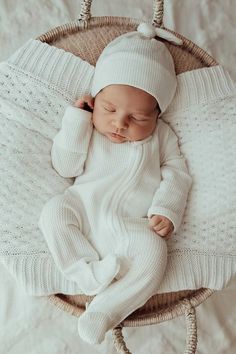 a baby in a white knitted outfit is laying on a wicker basket and wearing a white hat