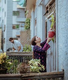 a woman holding a potted plant up to the side of a building while another man looks on