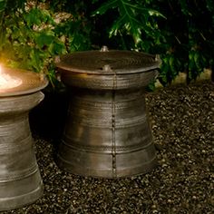 two metal candles sitting next to each other in front of some bushes and trees with green leaves