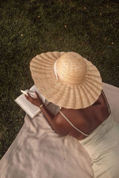 a woman in a white dress and straw hat reading a book on the grass with her hands behind her head