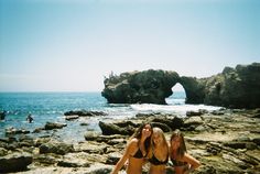 two women in bikinis standing on rocks near the ocean with an arch rock formation in the background