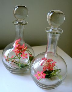 two glass vases with flowers in them on a white counter top, one is empty and the other has pink flowers inside