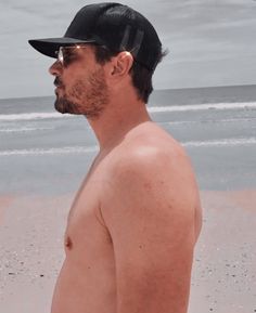 a shirtless man standing on the beach wearing sunglasses and a baseball cap with his hand in his pocket