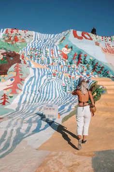 a woman is walking in front of a colorful wall with trees and mountains painted on it