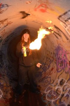 a woman standing in a tunnel holding a drink and fire stick with graffiti all over the walls