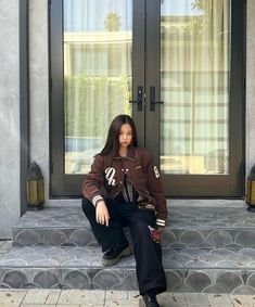 a woman is sitting on the steps in front of a door and wearing a brown jacket