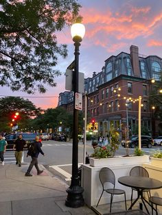 people are walking down the sidewalk in front of a building at dusk with lights on