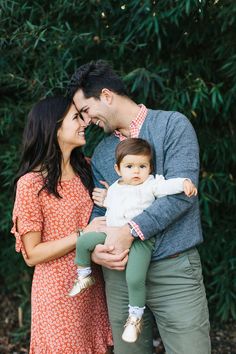 a man and woman holding a baby in front of some bushes with trees behind them