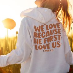 a woman wearing a white sweatshirt with the words we love because he first loves us printed on it