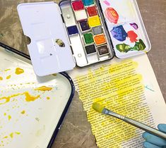 a person is painting with watercolors on an open book next to a paintbrush