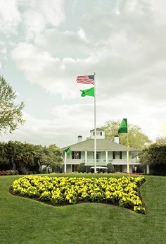 a flag flying in front of a white house with green and yellow flowers on the lawn