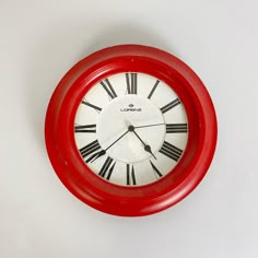 a red clock with roman numerals on the face is hanging on a white wall