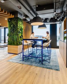 two people are sitting at a table in an office with plants on the wall behind them