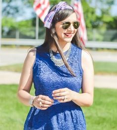 a woman wearing sunglasses and a blue dress is standing in the grass with an american flag behind her