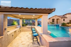 an outdoor kitchen and dining area next to a swimming pool at dusk with lights on