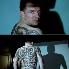 a man with tattoos on his back standing in front of a wall and looking at the camera