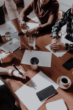 group of people sitting around a wooden table with papers and pens on it, looking at each other's hands