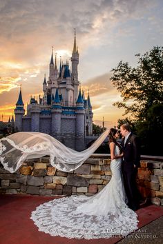 a bride and groom standing in front of a castle at sunset with their veil blowing in the wind