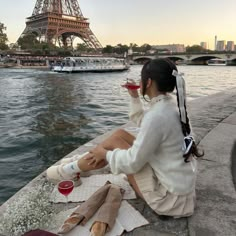 a woman sitting on the edge of a river drinking wine in front of the eiffel tower