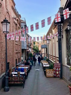 people are walking down an alley way with flags hanging from the buildings on either side