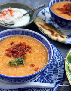 three bowls of soup on plates with spoons and other food items in the background