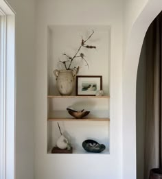 two shelves with vases and other items on them in a white walled room next to a window