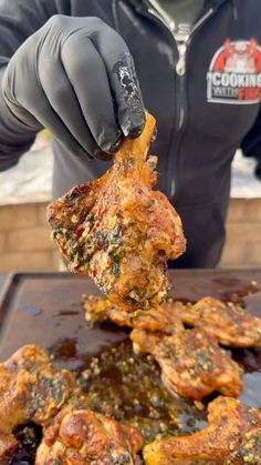 a person in black jacket and gloves holding up food over a pan with chicken on it