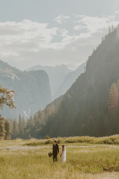 a bride and groom are walking through the grass with mountains in the background on their wedding day