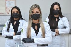 three women in white lab coats and black face masks standing next to each other with their arms crossed