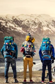 three men with backpacks standing on top of a mountain looking out at the snow covered mountains