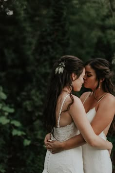 two women in white dresses standing next to each other with trees and bushes behind them