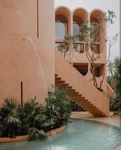 a fountain in front of a building with stairs and plants on either side of it