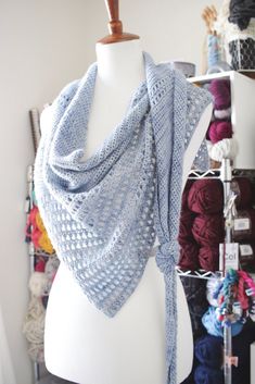 a white mannequin wearing a blue knitted shawl with multiple skeins of yarn in the background